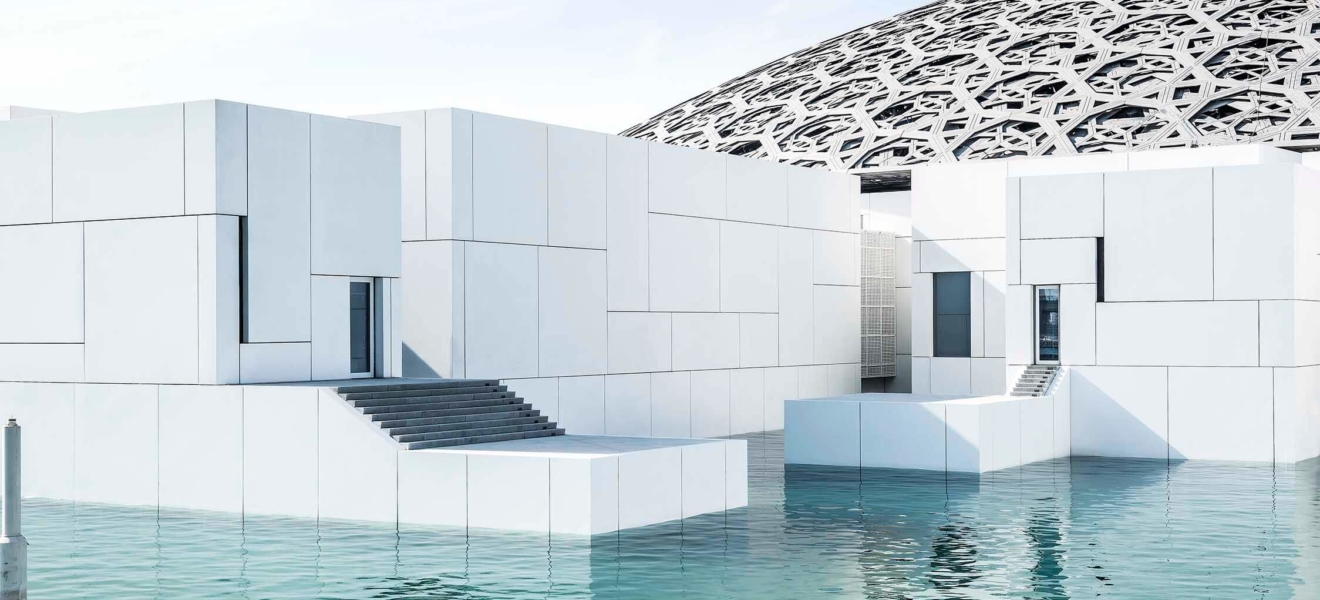 Aussenaufnahme vom Kunstmuseum Louvre von Jean Nouvel in Abu-Dhabi. Weisse Baukörper mit riesigem Kuppeldach, umgeben von türkisfarbenem Meerwasser.