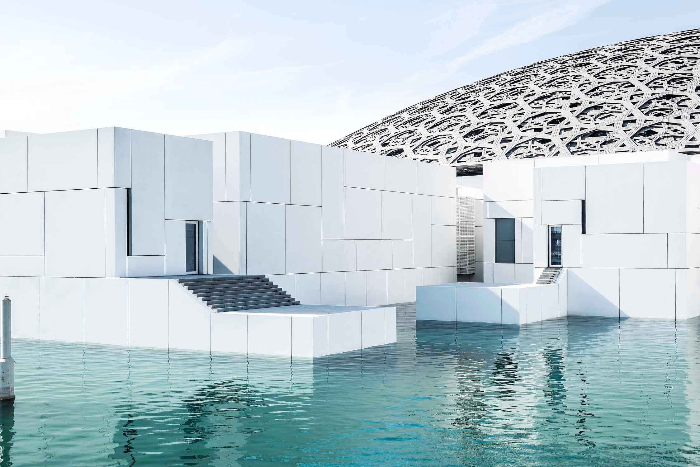 Aussenaufnahme vom Kunstmuseum Louvre von Jean Nouvel in Abu-Dhabi. Weisse Baukörper mit riesigem Kuppeldach, umgeben von türkisfarbenem Meerwasser. Architektur Fotografie Regine Giesecke Architekturfotograf Architecture Photography