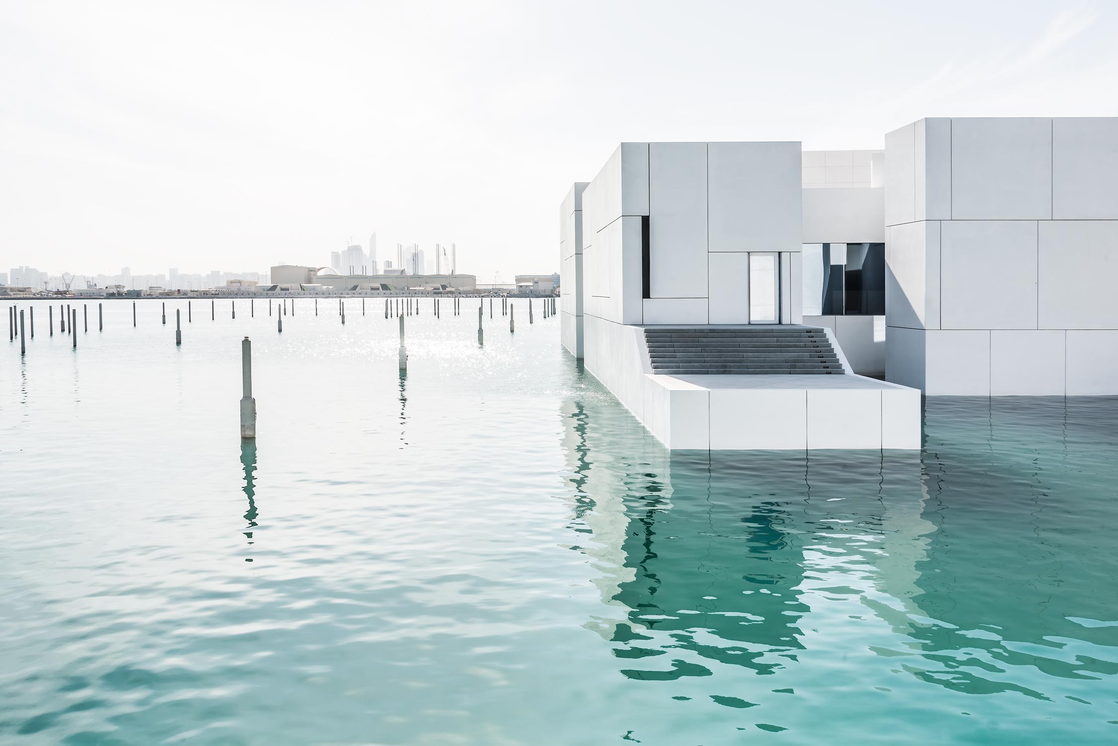 Aussenaufnahme vom Kunstmuseum Louvre von Jean Nouvel in Abu-Dhabi. Weisse Baukörper mit riesigem Kuppeldach, umgeben von türkisfarbenem Meerwasser.