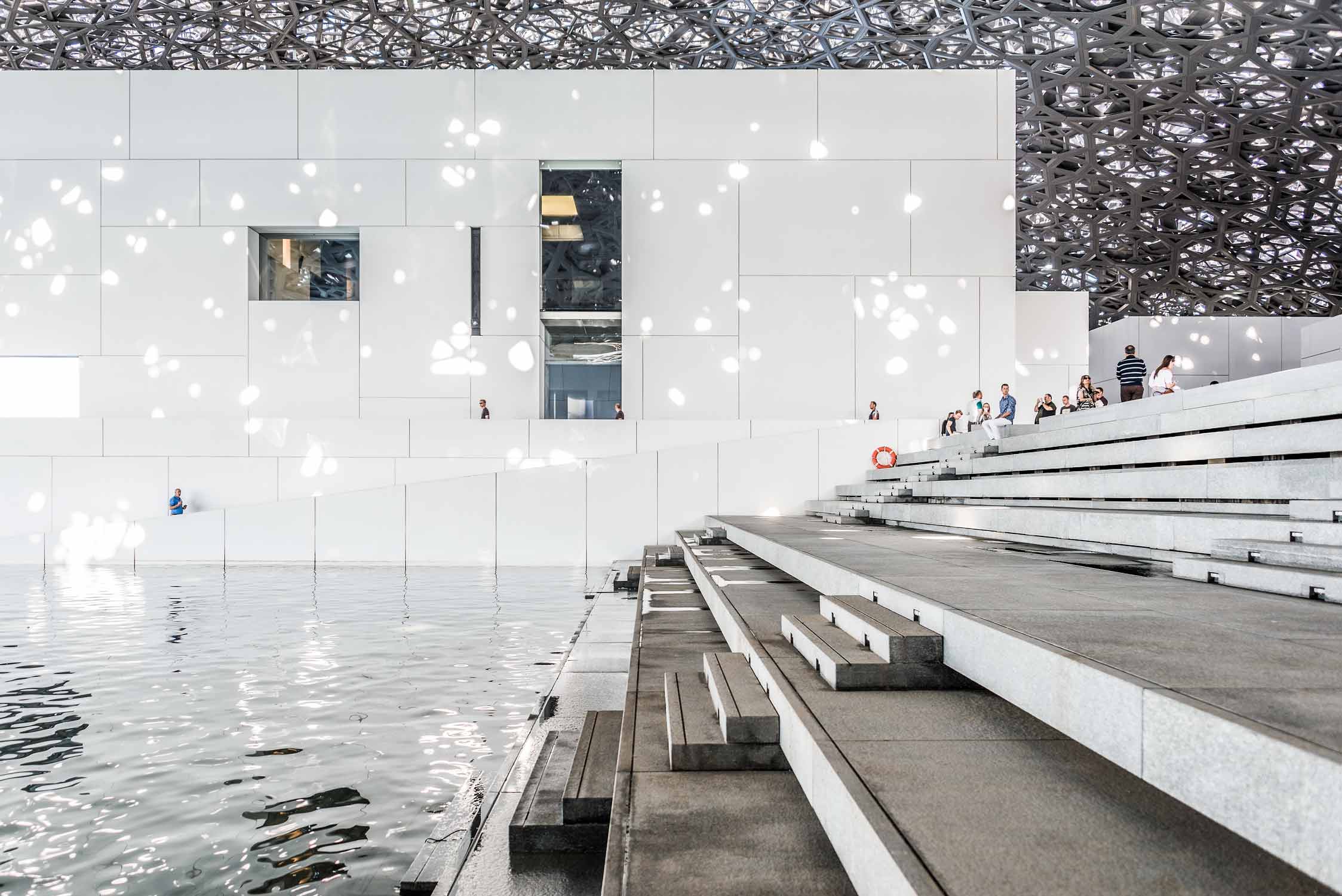 Anlegestelle mit Sitzstufen im Kunstmuseum Louvre von Jean Nouvel in Abu-Dhabi. Weisse Baukörper mit riesigem Kuppeldach, das Lichtstrahlen, wie einen Lichtregen durchlässt.
